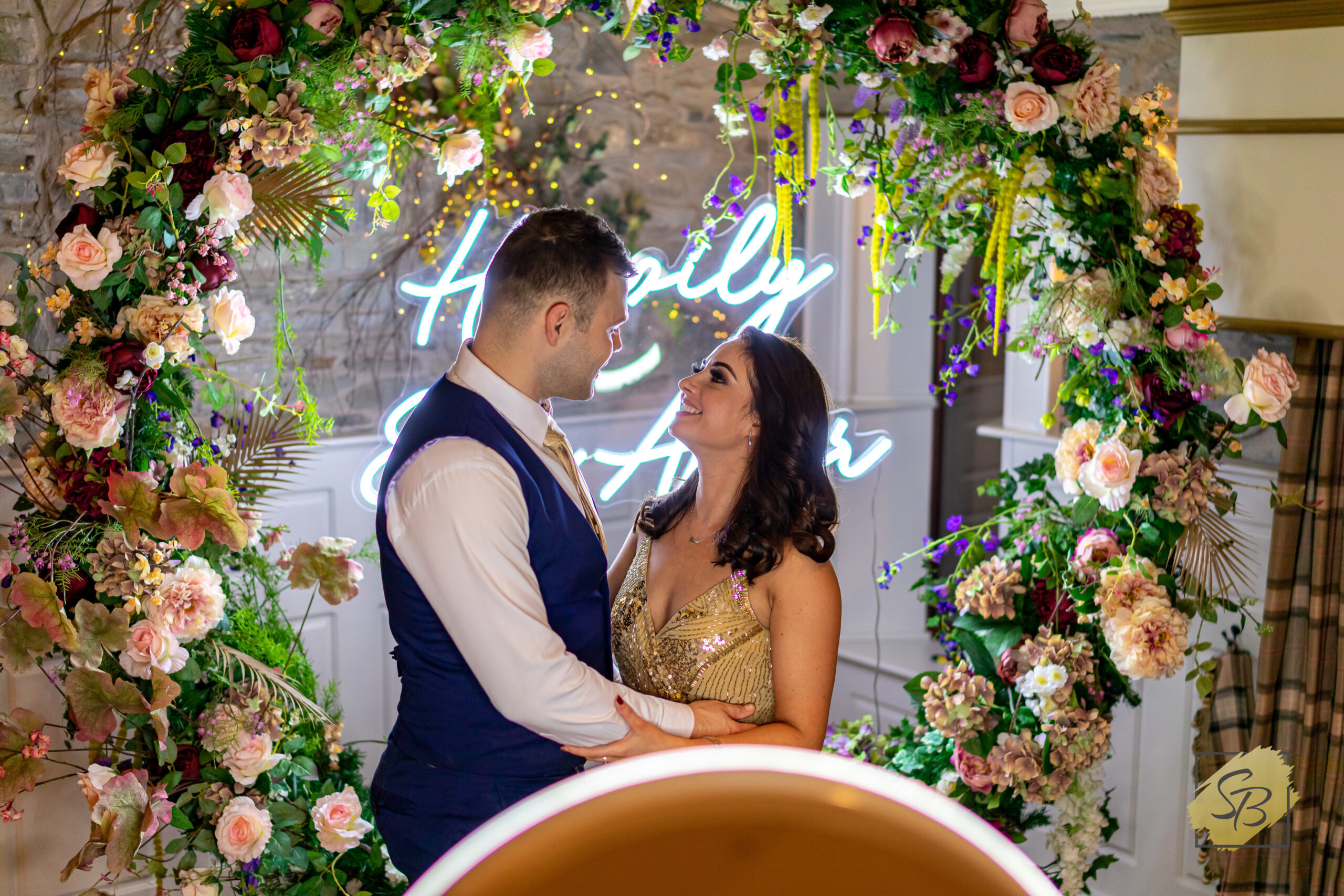 A beautiful couple of wedding guests posing for photos to be taken on the Mobi Aura, in front of the flower ring backdrop and the happily ever after neon sign
